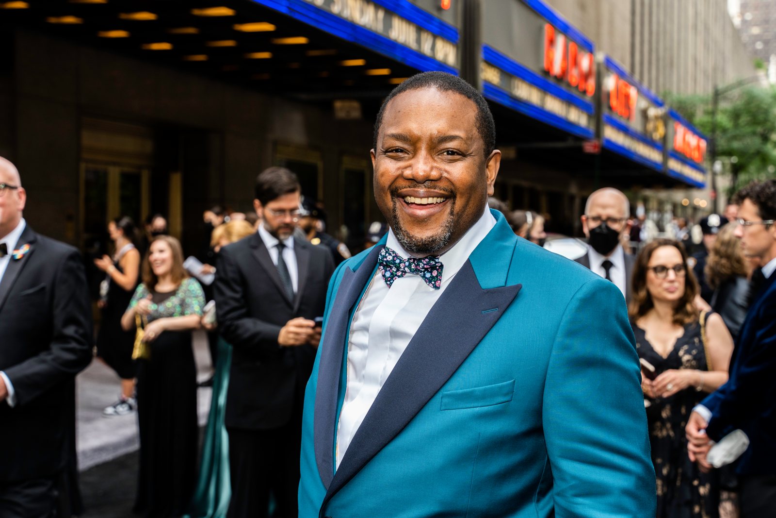 Smiling red carpet portrait in a teal tuxedo jacket and floral bow tie outside Radio City at the Tony Awards